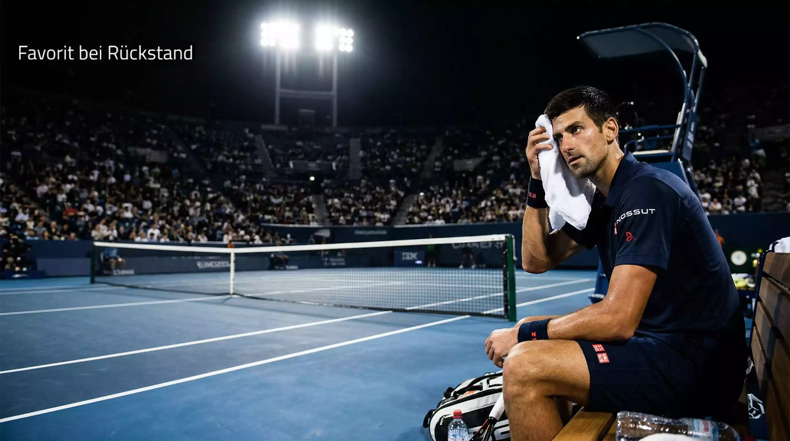 Tennisspieler wischt sich Schweiß ab und blickt entschlossen auf den Platz bei Flutlicht – Favorit bei Rückstand